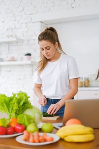A young woman in a white t-shirt chops fresh vegetables on a wooden board in a bright kitchen.