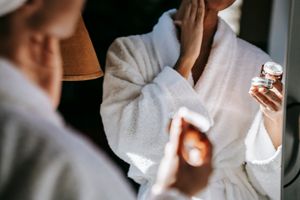 Crop anonymous female in white bathrobe applying moisturizing facial cream on face while standing in front of mirror in bathroom