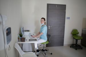 Side view of young slim female medic in uniform and face mask typing on keyboard while using computer and sitting on chair in modern hospital office and looking at camera